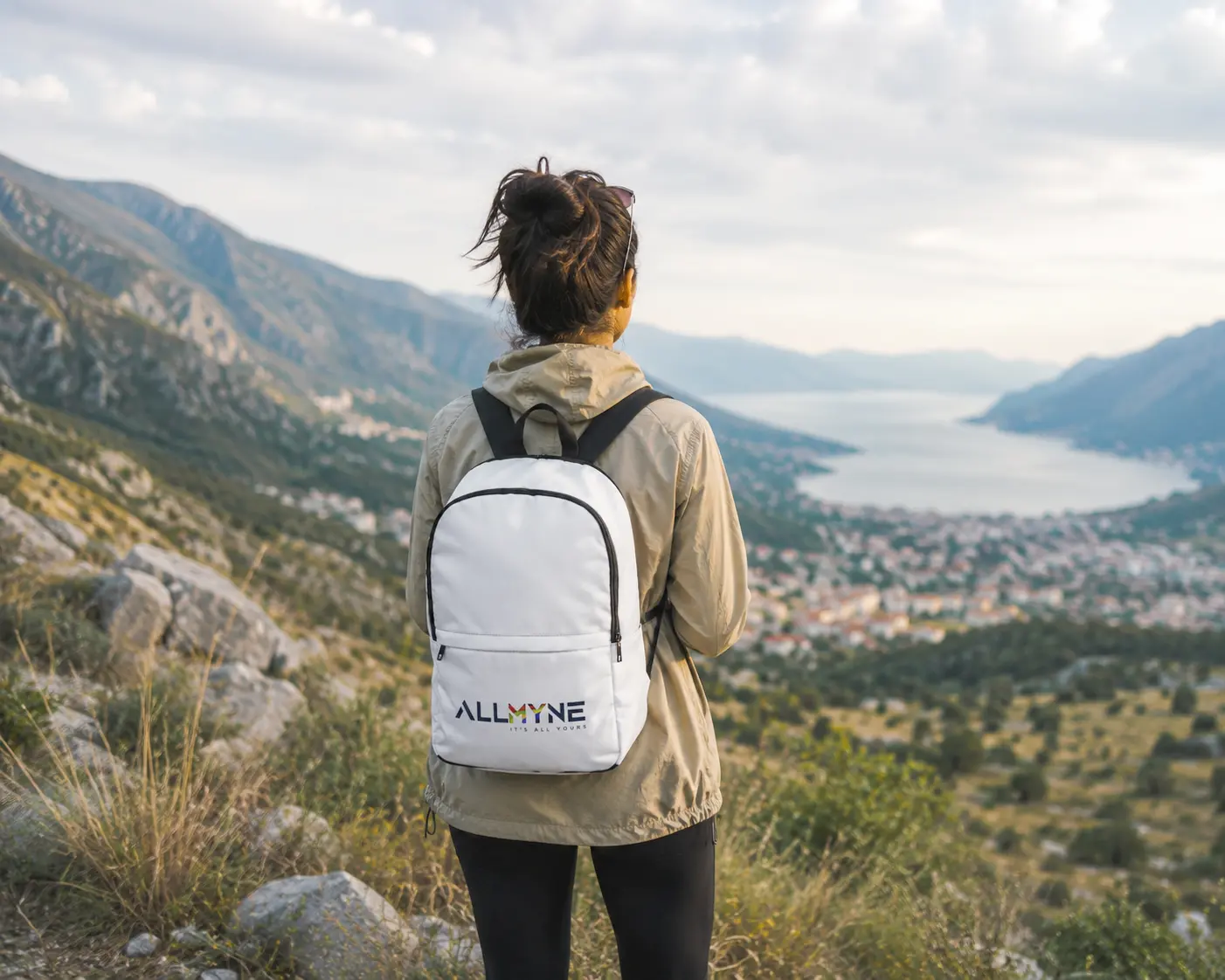 Solo traveler wearing an ALLMYNE backpack standing on a mountain ridge overlooking a scenic coastal valley.